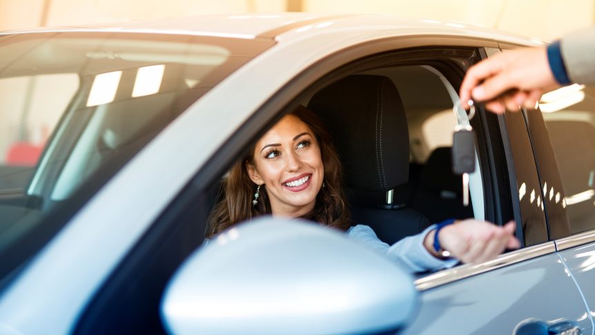 An attractive brunette woman sitting in her brand new car and taking keys from vehicle dealer.